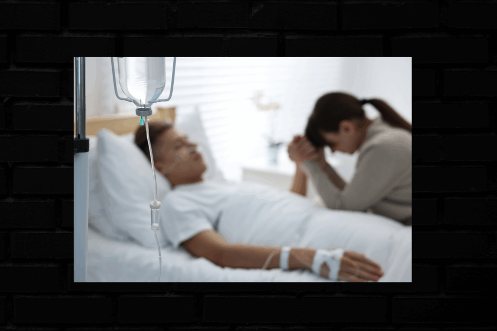 A woman prays in a hospital room beside a man with an IV in his arm.