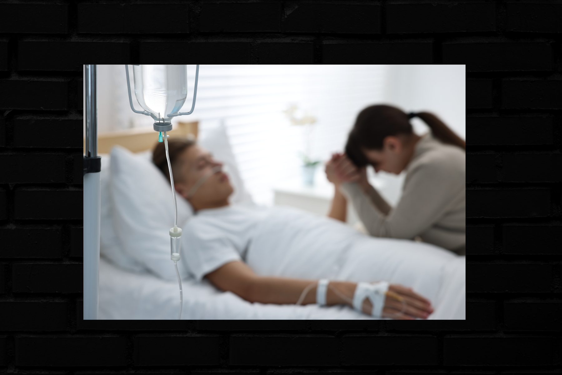 A woman prays in a hospital room beside a man with an IV in his arm.